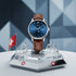 Watch with brown leather strap on a stand featuring Swiss flag design against a geometric background