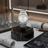 Silver watch on a black marble stand with architectural models and plans in the background
