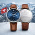 Two watches with brown leather straps backcase engraved in front of a mountainous landscape with a Swiss flag.